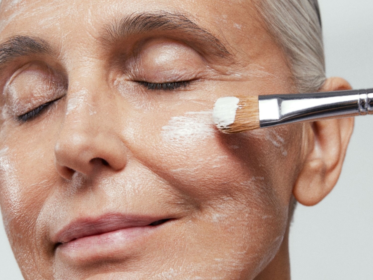 Nahaufnahme einer Frau mit geschlossenen Augen, deren Gesicht mit einer weißen Creme bedeckt ist, die mit einem Pinsel aufgetragen wird.
