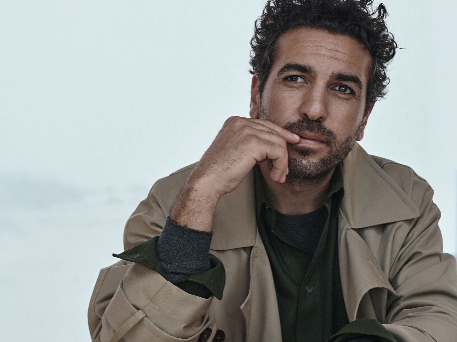 Ein Mann mit lockigem, dunklem Haar und Bart trägt einen beigen Trenchcoat über einem grünen Hemd und einem grauen Unterhemd, während er nachdenklich zur Seite blickt.
