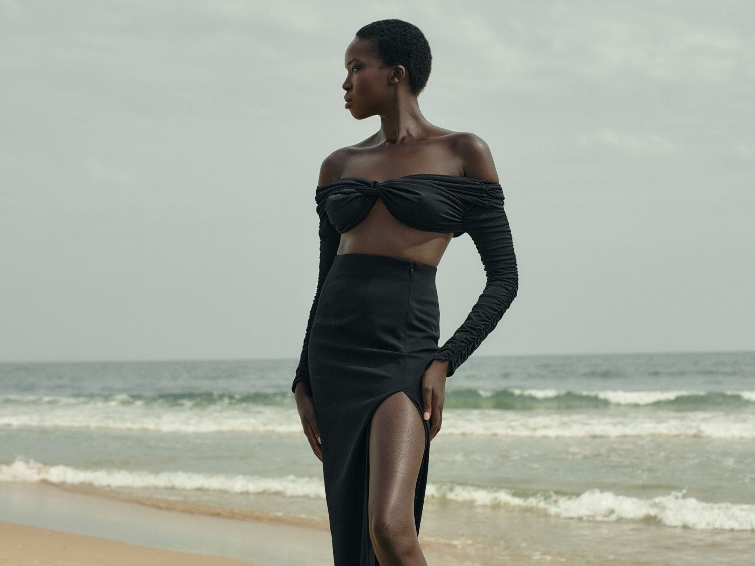 Eine Frau mit dunkler Haut steht am Strand und trägt ein schwarzes, schulterfreies Oberteil mit langen Ärmeln und einen Rock mit Schlitz.