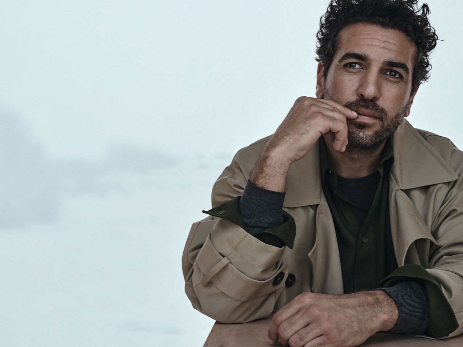 Ein Mann mit lockigem, dunklem Haar und Bart trägt einen beigen Trenchcoat über einem grünen Hemd und einem grauen Pullover, während er nachdenklich mit der Hand am Kinn sitzt.