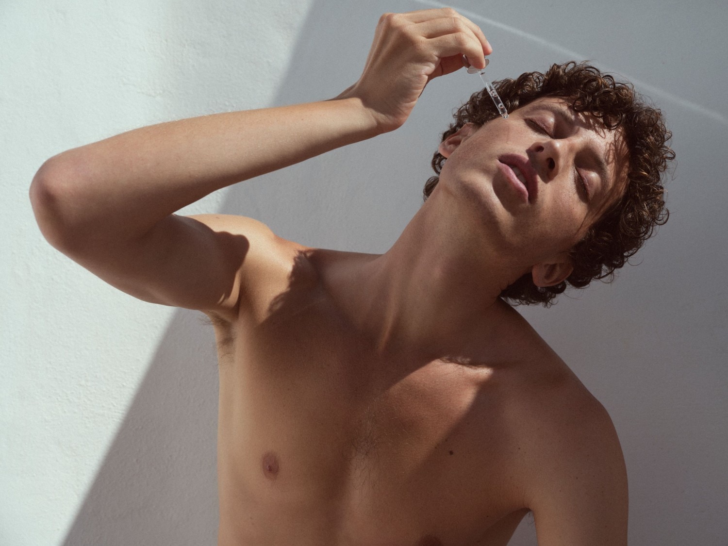 Jaunuolis užsimerkęs lašina skystį iš pipetės ant veido, pabrėždamas odos priežiūros svarbą.