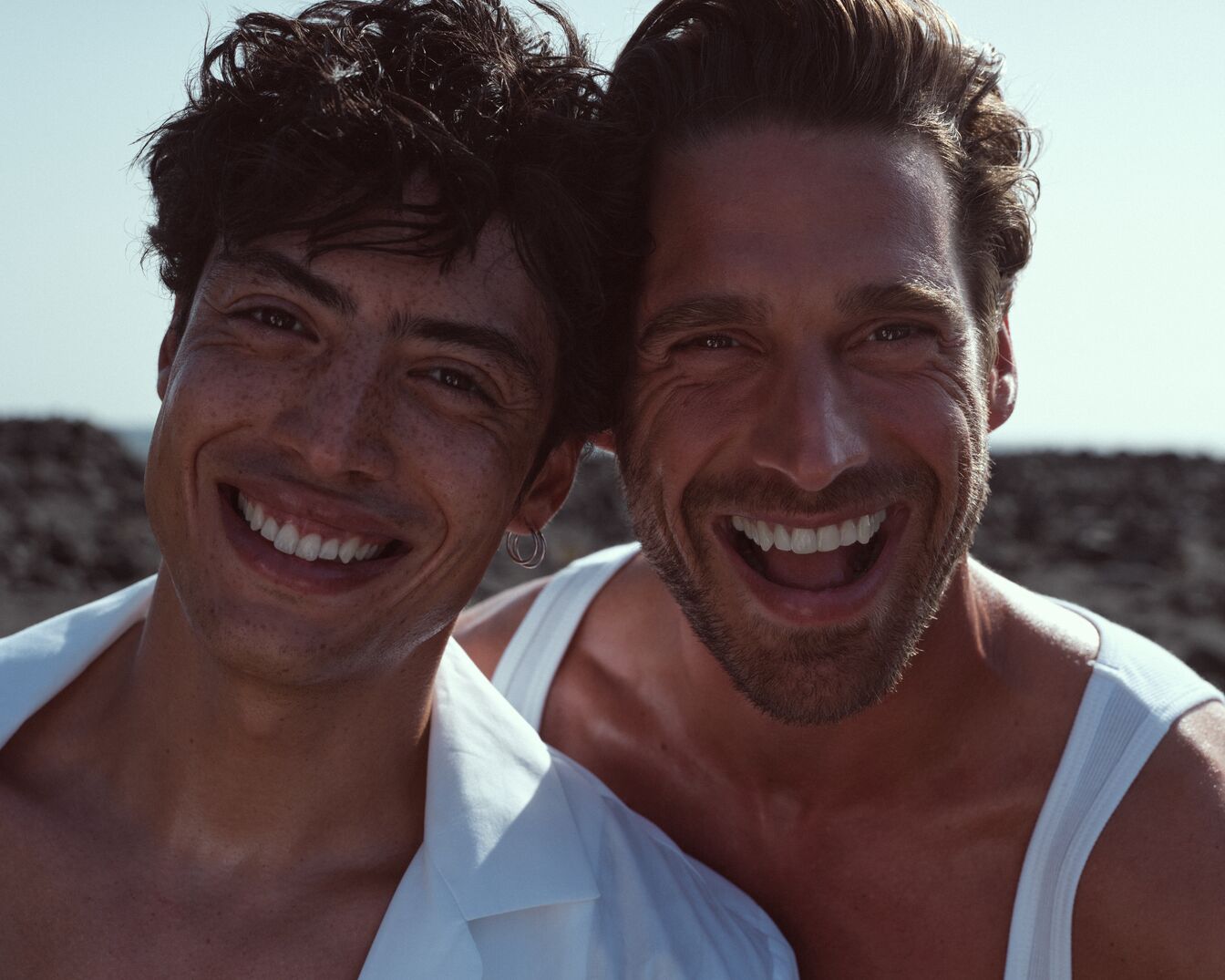 Nahaufnahme von zwei Männern, die eng beieinander stehen und breit lächeln, wobei der eine ein weißes Hemd und der andere ein weißes Tanktop trägt.