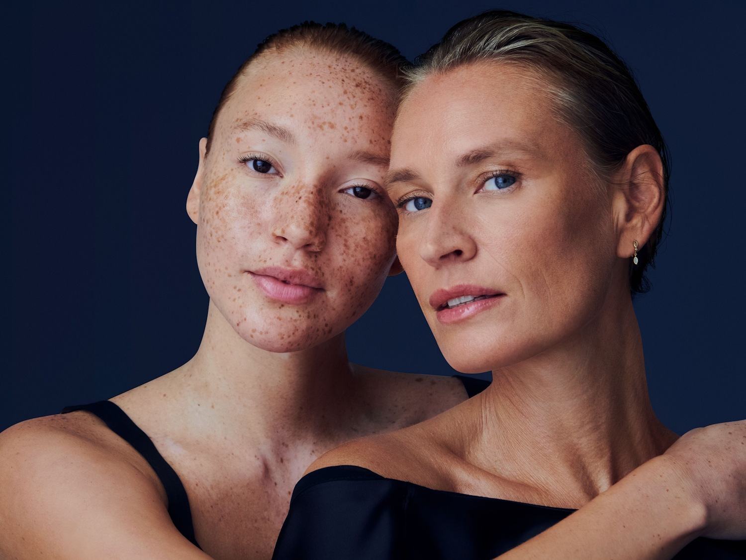 Švelnus dviejų moterų portretas tamsiai mėlyname fone: viena su strazdanotu veidu ir juodais marškinėliais, kita – su šviesiais plaukais ir subtiliais auskarais.