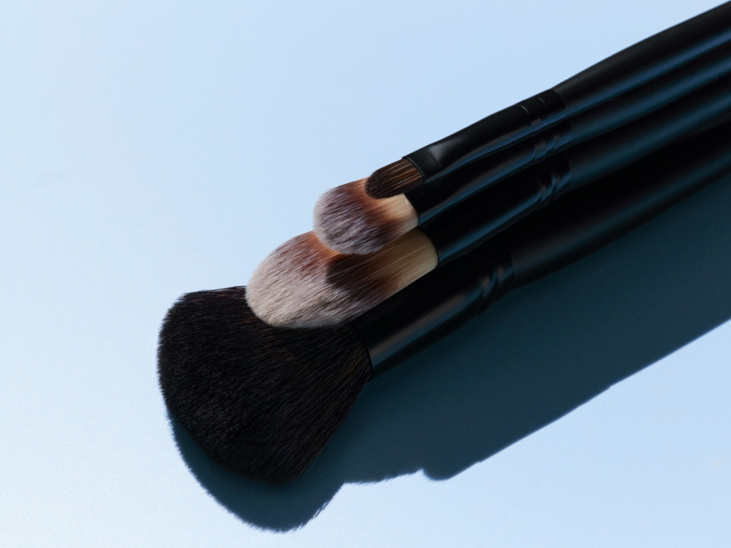 Trys makiažo šepetėliai su juodomis rankenomis ir rudais šereliais išdėstyti ant šviesiai mėlyno paviršiaus.