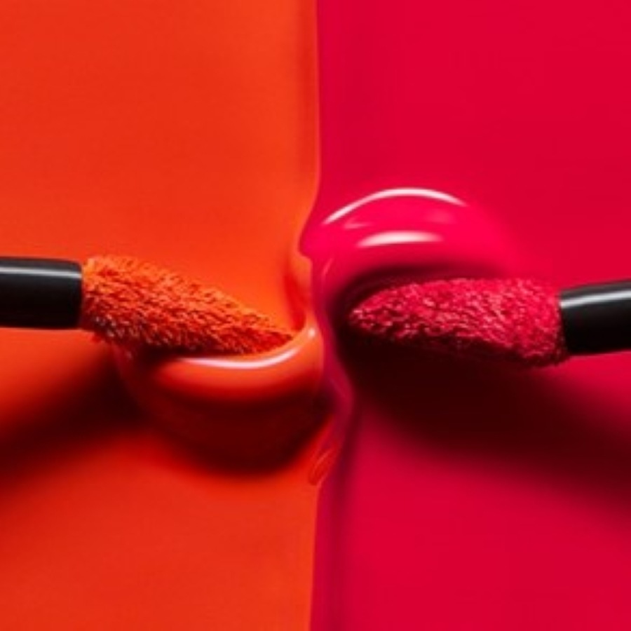 Nahaufnahme von zwei Lippenstiftfarben, Orange und Rot, mit Applikatorbürsten, die in die cremige Textur eintauchen.