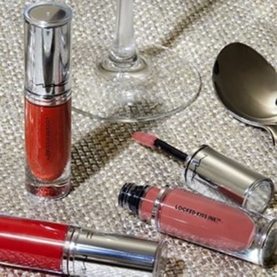 Nahaufnahme von MAC Locked Kiss Ink Liquid Lippenstiften in verschiedenen Farbtönen, die auf einem glitzernden Untergrund mit einem Glas und einem Löffel arrangiert sind.