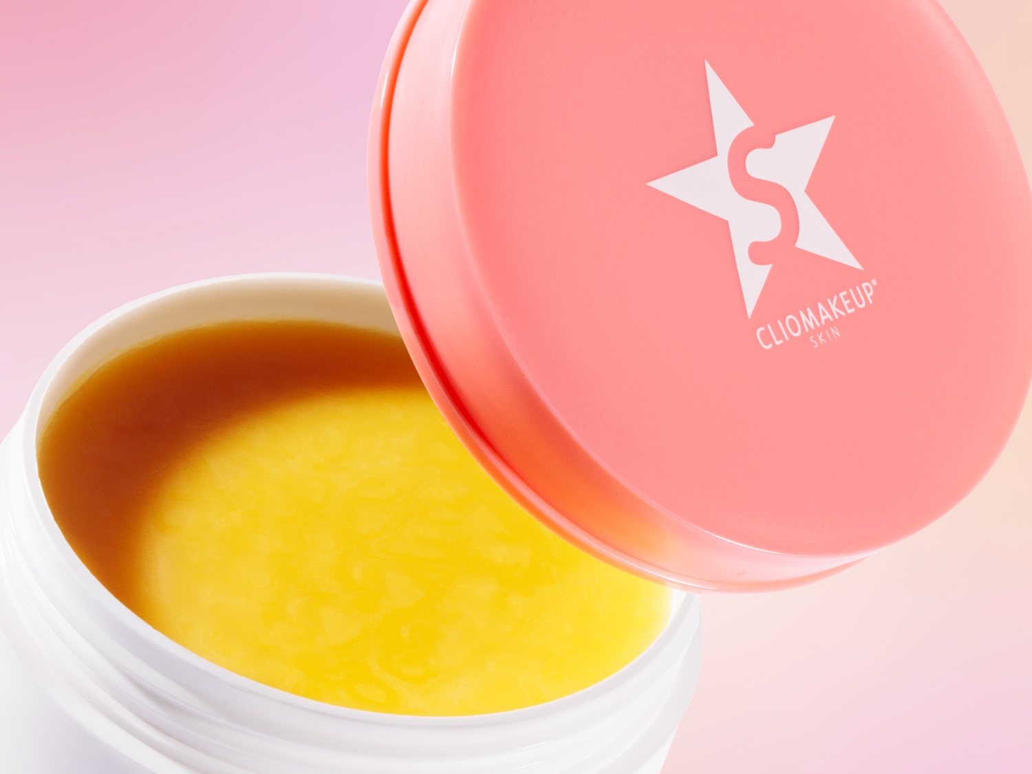 Ecco un barattolo bianco aperto di CLIO MAKEUP skin con un coperchio rosa chiaro che mostra il logo del marchio, che rivela una crema gialla liscia all'interno.