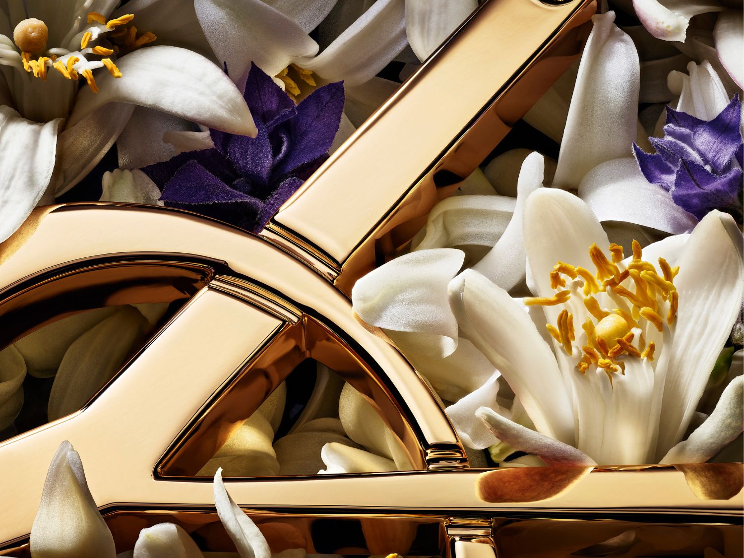 Detailný záber na zlaté logo YSL obklopené bielymi a fialovými kvetmi, vytvárajúci luxusný a elegantný dojem.