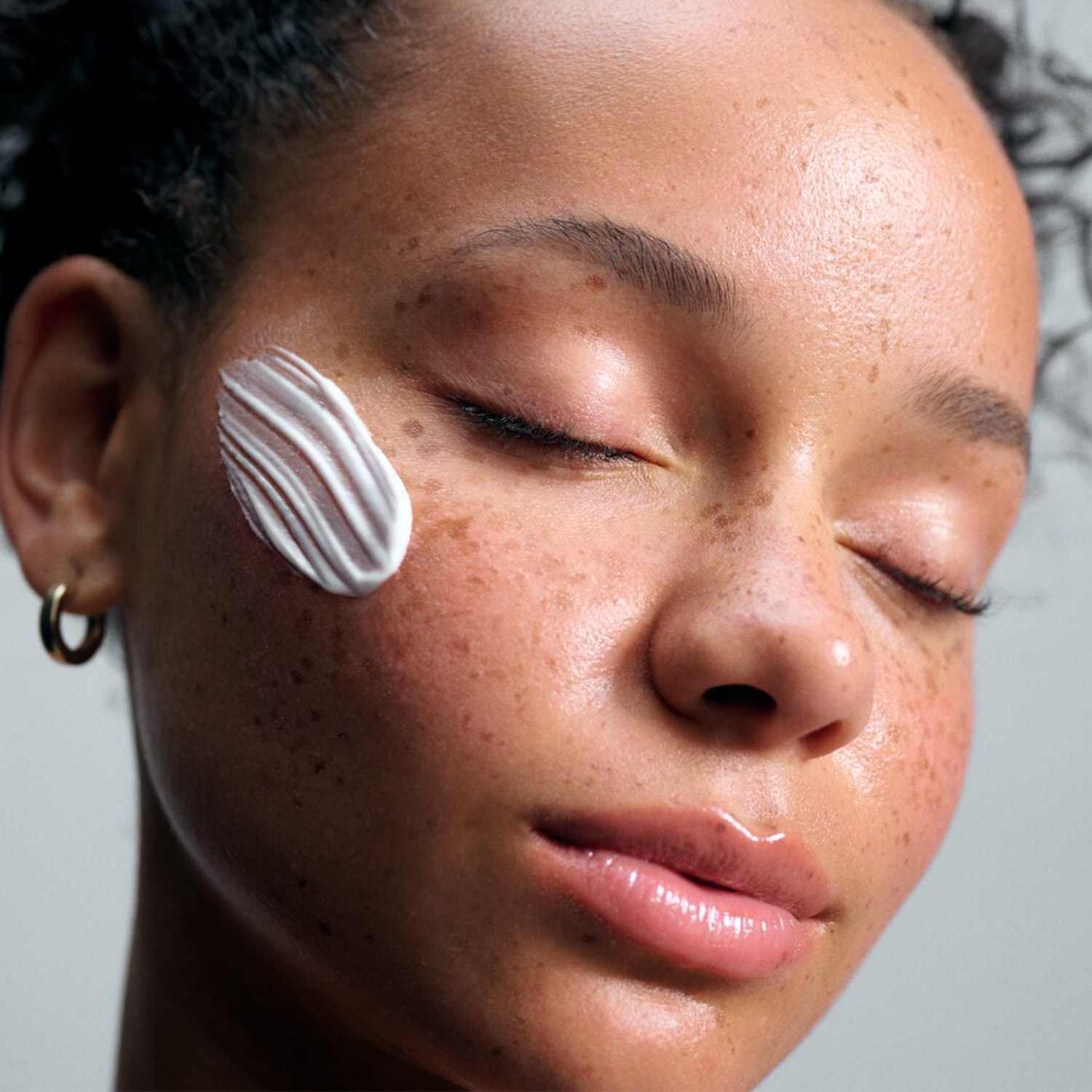 Een vrouw met gesloten ogen heeft een streep witte crème op haar wang, wat de hydratatie en verzorging van de huid benadrukt.