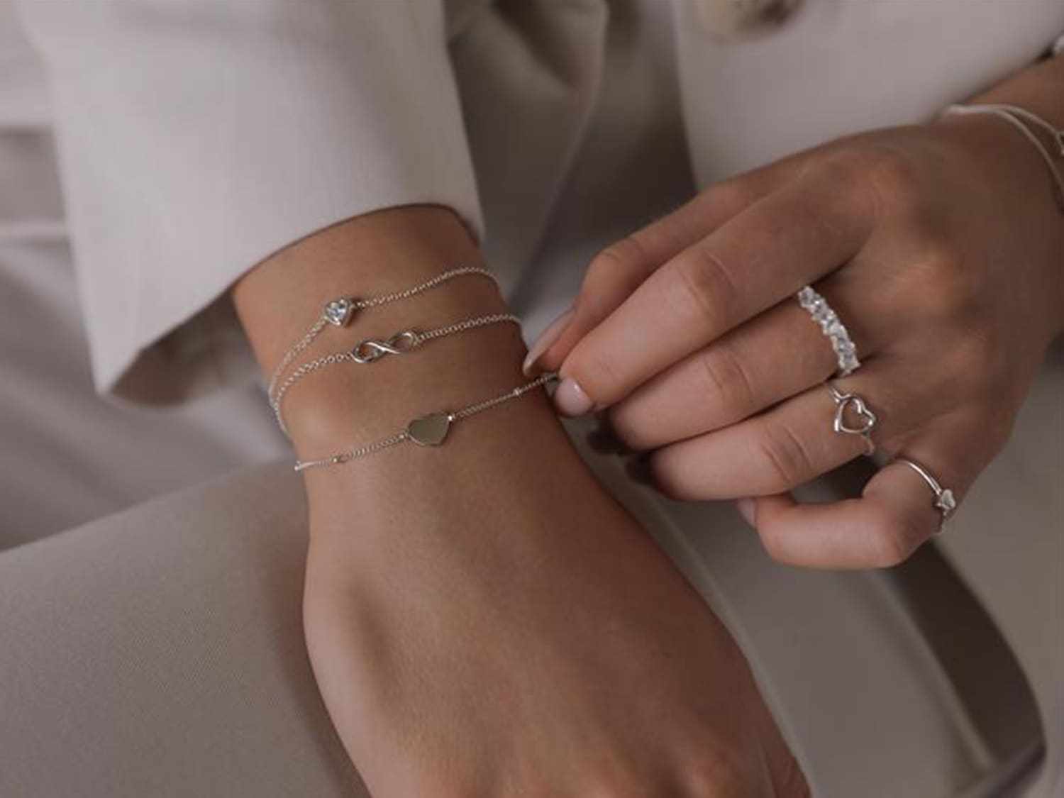 Nahaufnahme einer Hand, die mit drei silbernen Armbändern mit Herz-, Unendlichkeits- und Diamantanhängern sowie mehreren Ringen mit Herz- und Diamantdetails geschmückt ist.
