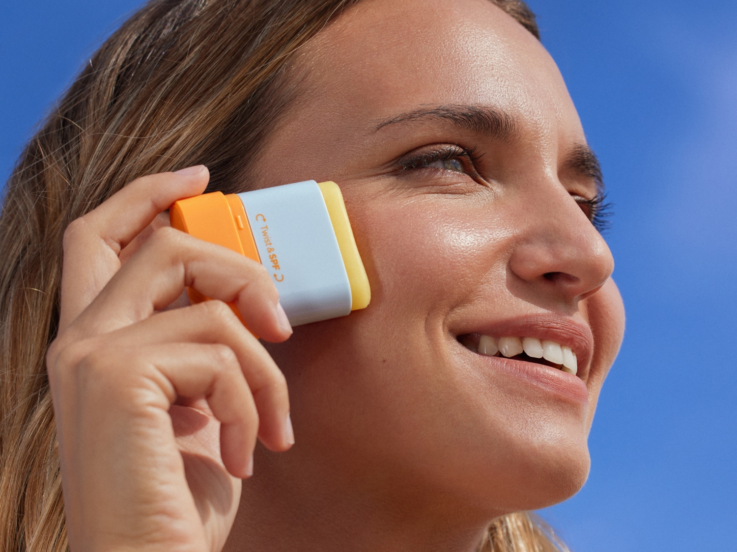 Una mujer sonriente aplica un protector solar en barra Twist & SPF de color blanco, amarillo y naranja en su mejilla, protegiendo su piel del sol.