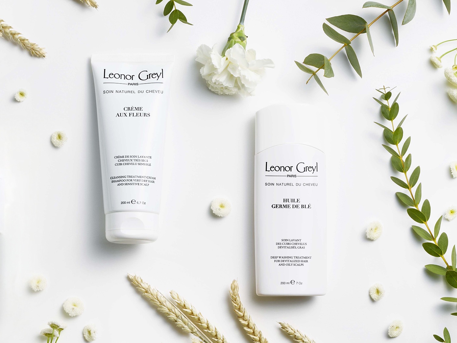 Na białym tle leżą dwa białe opakowania produktów Leonor Greyl do pielęgnacji włosów, otoczone kwiatami i zielonymi gałązkami.