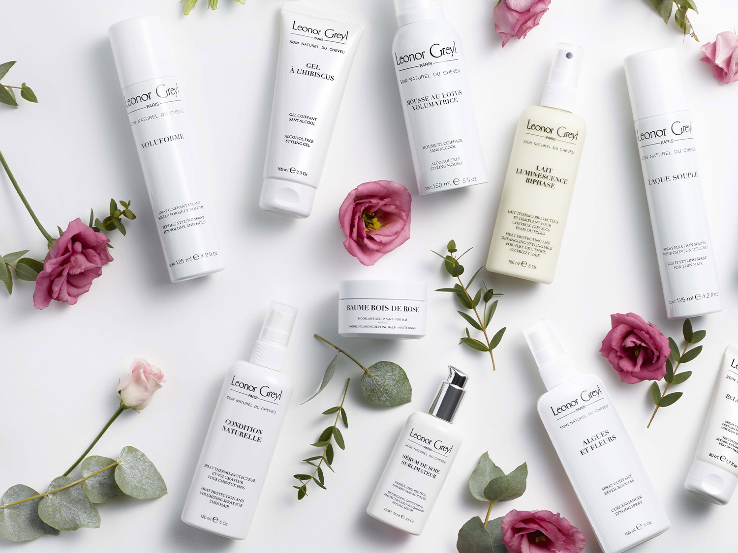 Na białym tle leżą produkty do włosów Leonor Greyl w białych opakowaniach, otoczone różowymi kwiatami i zielonymi listkami, tworząc elegancką kompozycję.