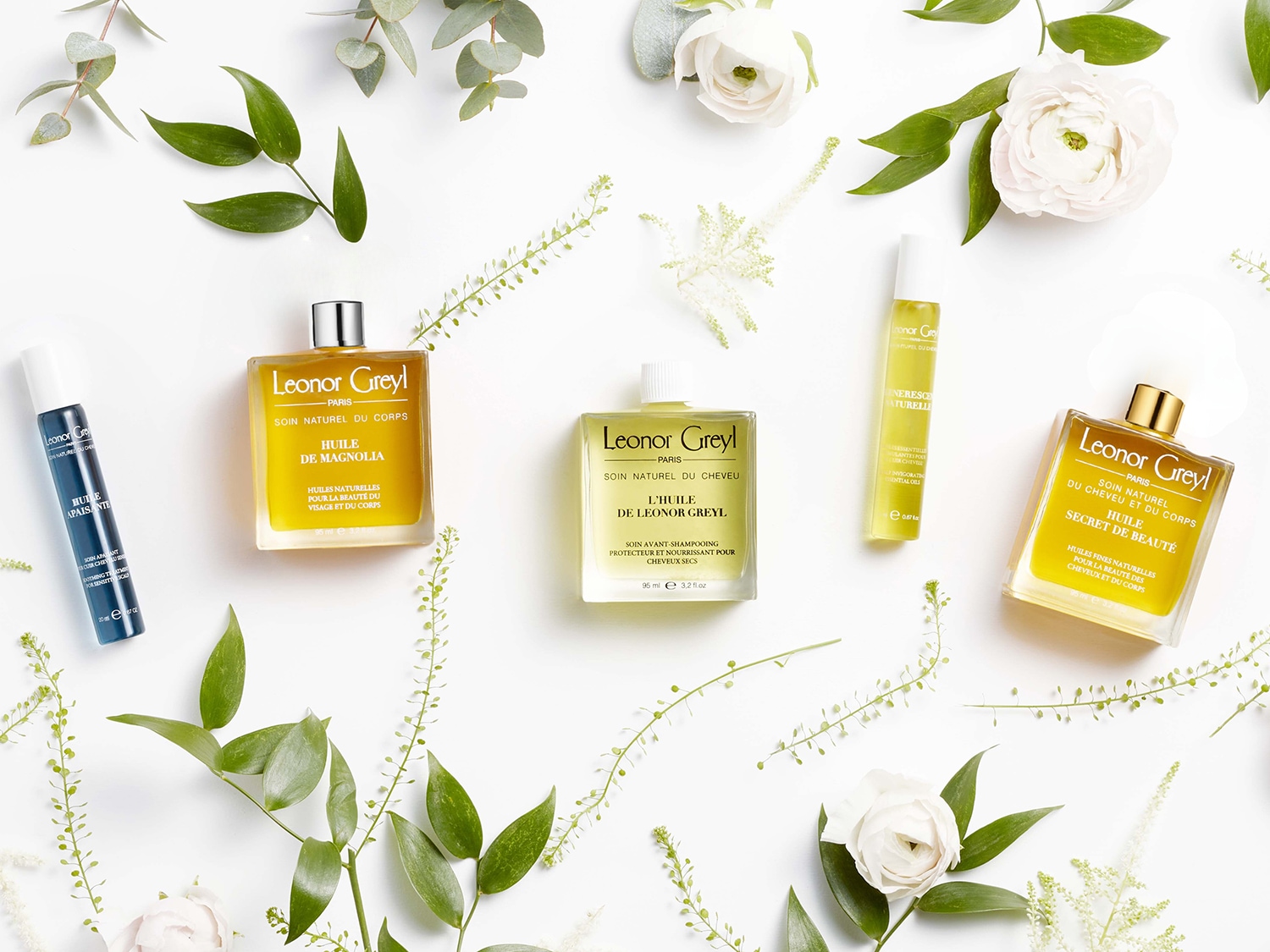 Na białym tle leżą produkty Leonor Greyl, w tym olejki do ciała i włosów, otoczone białymi kwiatami i zielonymi liśćmi, tworząc naturalną kompozycję.