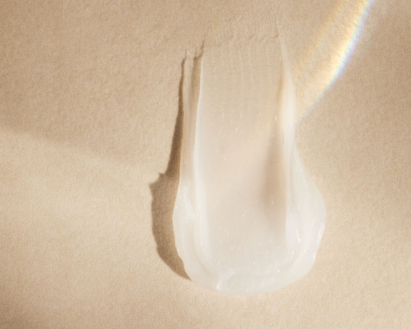 Een close-up toont een veeg doorschijnende, lichtbeige crème met een glanzende textuur op een beige achtergrond.