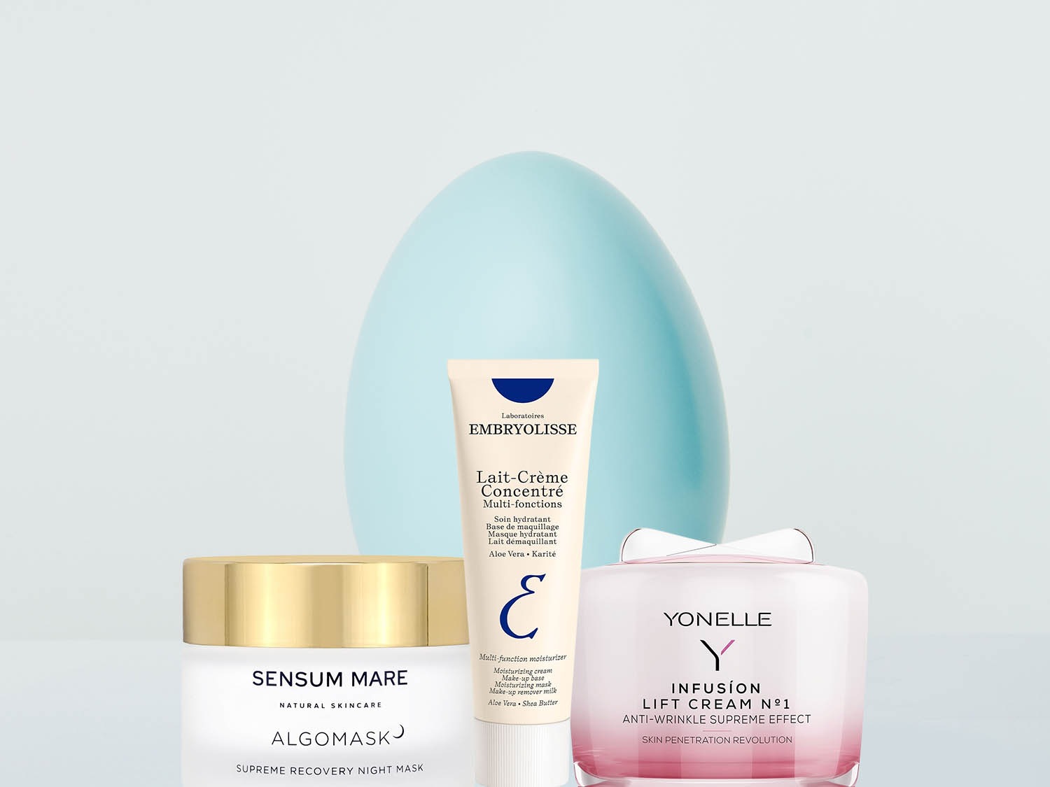 Na pierwszym planie trzy kremy do twarzy: Sensum Mare Algomask w białym słoiczku ze złotą nakrętką, Embryolisse Lait-Crème Concentré w tubce i Yonelle Infusion Lift Cream N°1 w różowym słoiczku.