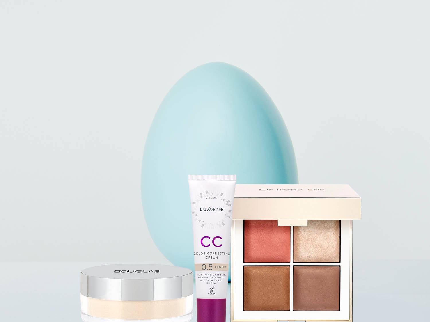 Kompaktowy puder Douglas, krem CC Lumene w białej tubce z fioletowymi akcentami, paleta do makijażu Dr Irena Eris z czterema odcieniami oraz dekoracyjne, błękitne jajo w tle.