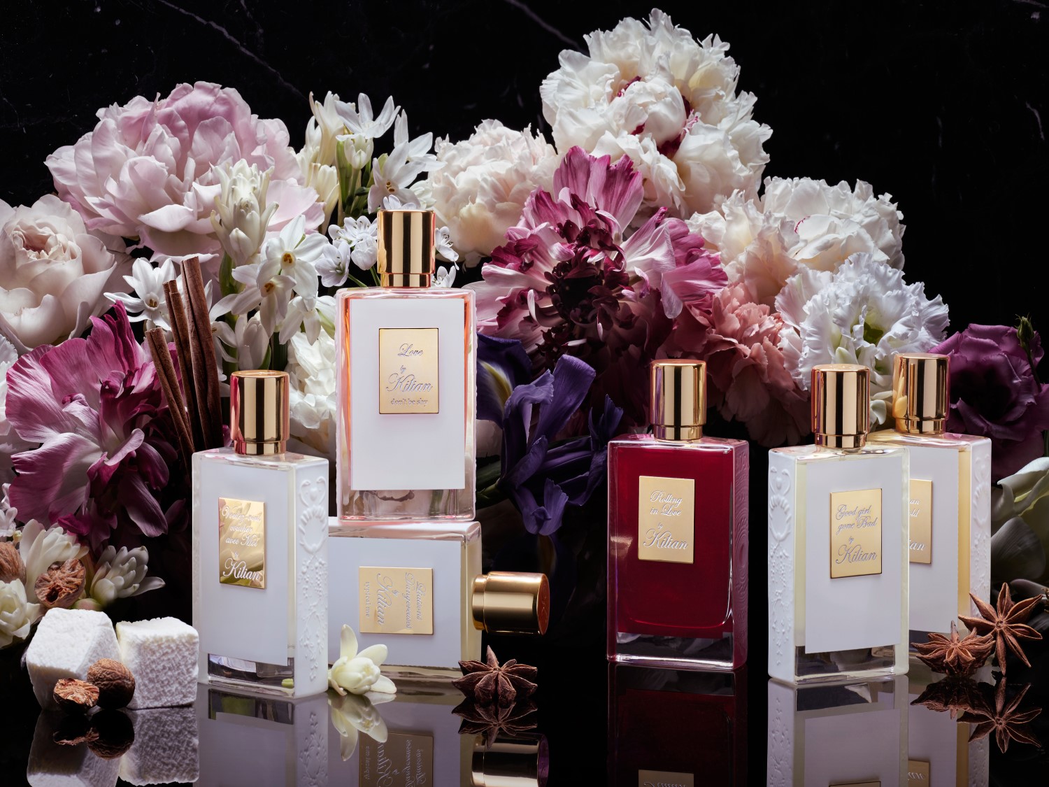 Elegancka kolekcja zapachów Kilian w białych i czerwonych flakonach ze złotymi korkami, otoczona kwiatami w odcieniach różu i bieli, tworzy luksusową kompozycję.