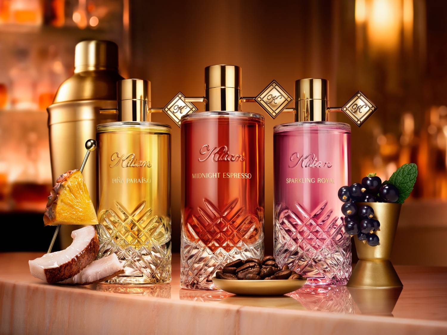 Trzy butelki perfum Kilian Paris, Piña Paraíso, Midnight Espresso i Sparkling Royal, stoją na marmurowym blacie, otoczone składnikami, które odzwierciedlają ich zapachy.