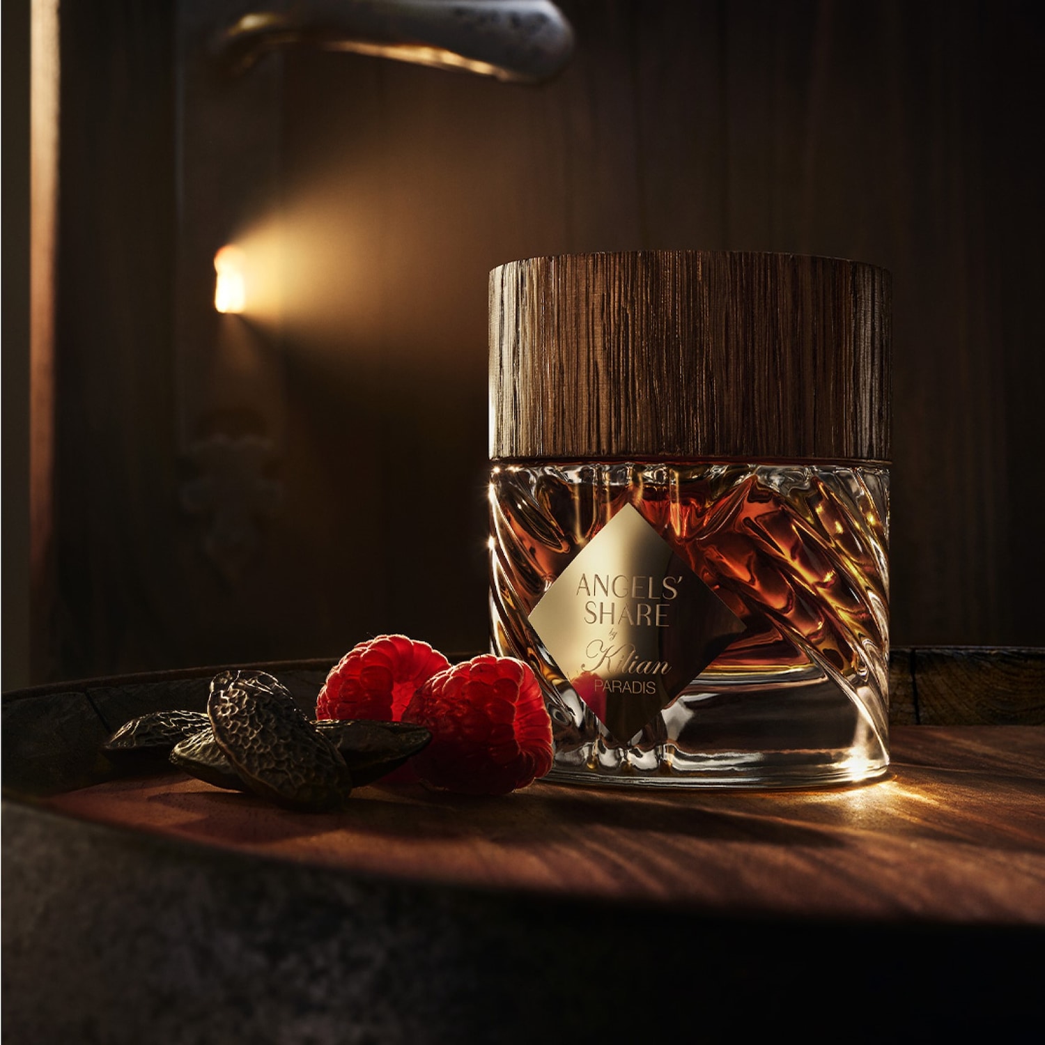 Butelka bursztynowego płynu Kilian Angels' Share Paradis stoi na drewnianej powierzchni obok malin i ciemnych ziaren, z drewnianym korkiem i złotą etykietą.