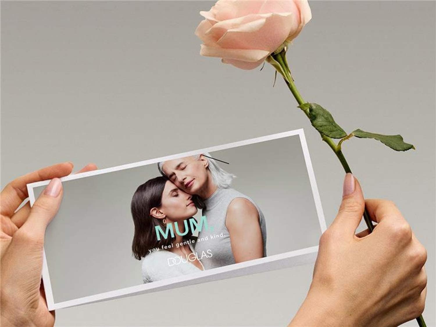 Une main tient une carte de Douglas avec une photo d'une mère et de sa fille, avec le texte "Maman, tu te sens douce et gentille", à côté d'une rose rose.