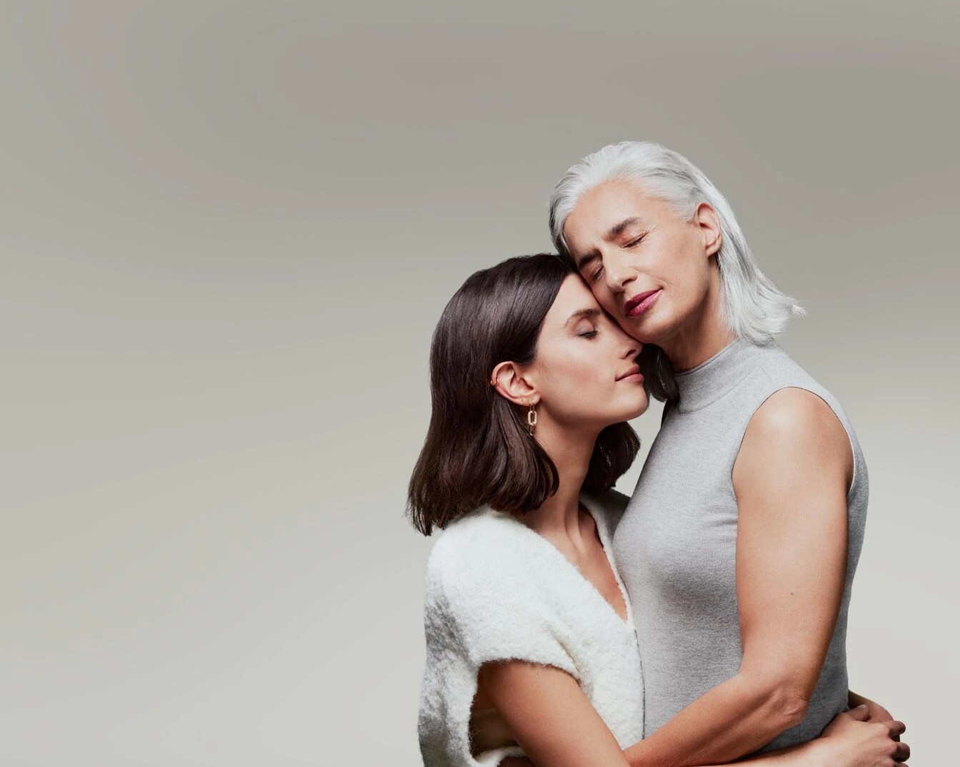 Une jeune femme aux cheveux bruns et une femme plus âgée aux cheveux gris se tiennent enlacées, les yeux fermés, dans un moment de tendresse.