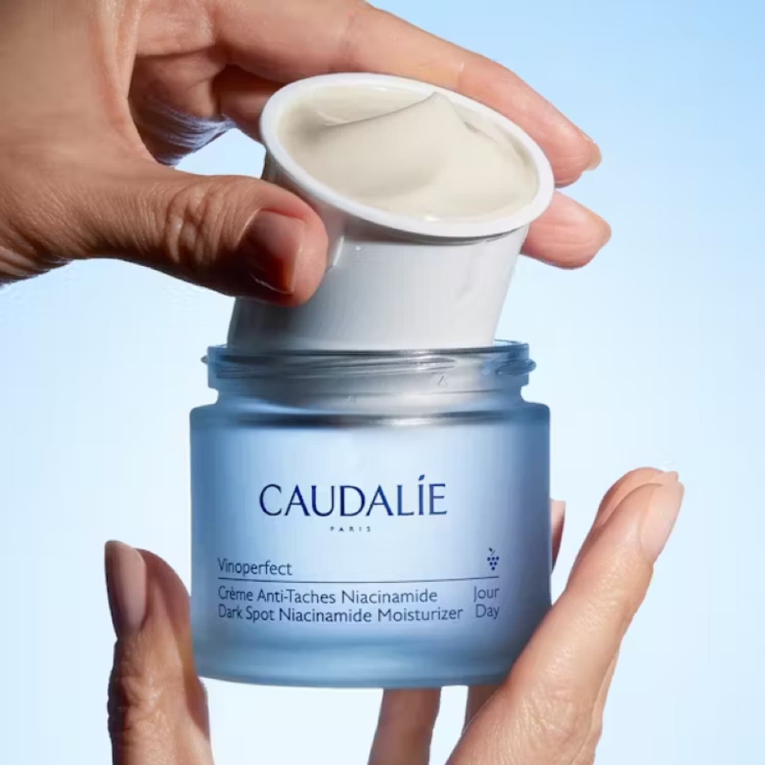 Na dłoniach prezentuje się słoiczek Caudalie Vinoperfect, krem na dzień redukujący przebarwienia z niacynamidem, z otwartym wieczkiem, ukazującym kremową konsystencję.