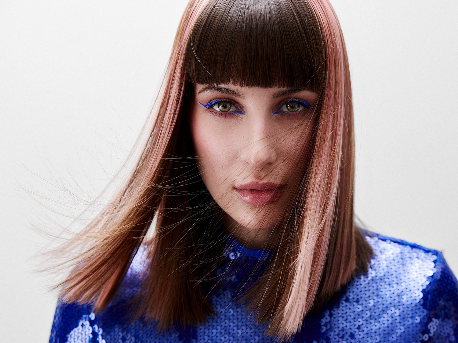 Moters portretas su ryškiai mėlynu akių kontūru, tamsiai rudais plaukais ir rožiniais akcentais, vilkinti mėlyną blizgučių palaidinę.