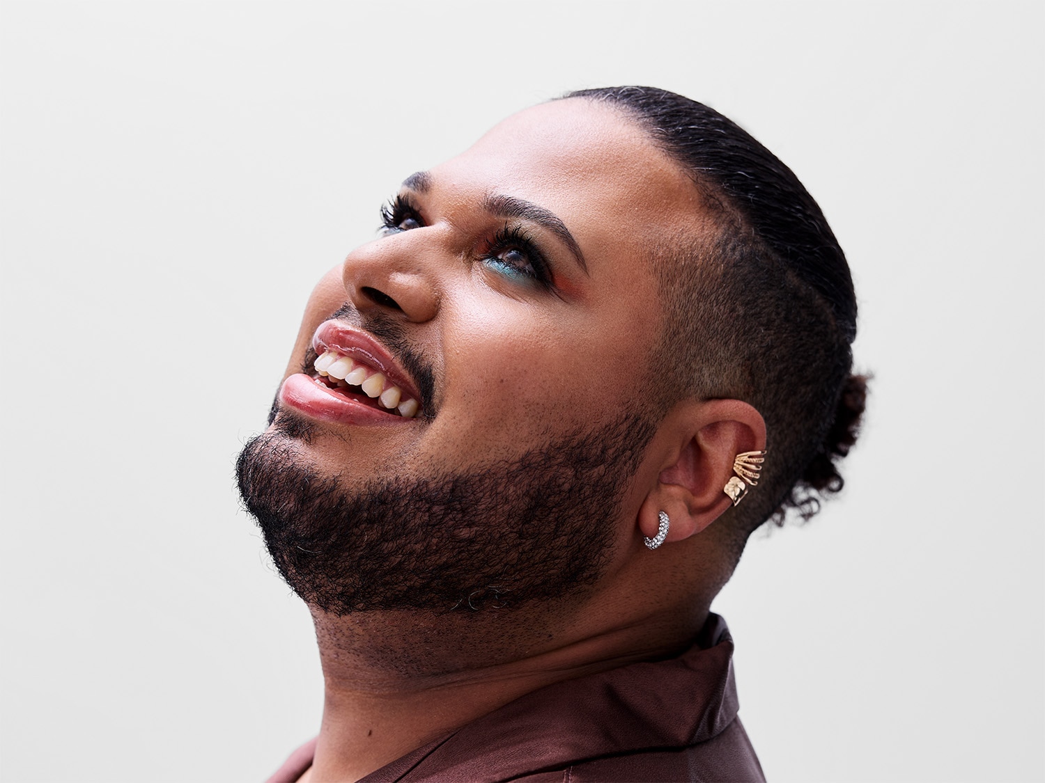 Švytintis vyras su ryškiu makiažu, įskaitant blizgančius lūpų dažus ir žydrus akių šešėlius, žiūri aukštyn su šypsena, demonstruodamas savo unikalų stilių ir džiaugsmą.