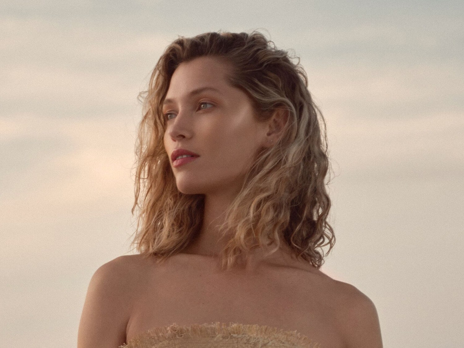 Primo piano di una donna con capelli biondi ondulati e labbra rosate, che guarda verso sinistra con un'espressione serena.