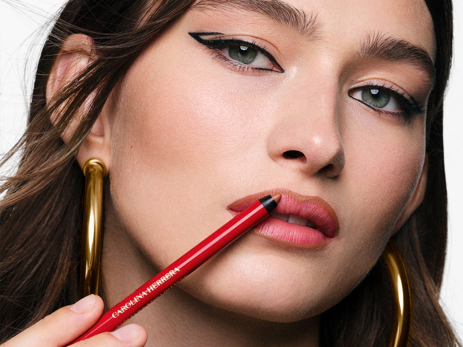 Nahaufnahme eines Gesichts mit einem roten Carolina Herrera Lipliner, der auf die Lippen aufgetragen wird, dazu goldene Ohrringe und ein Lidstrich.