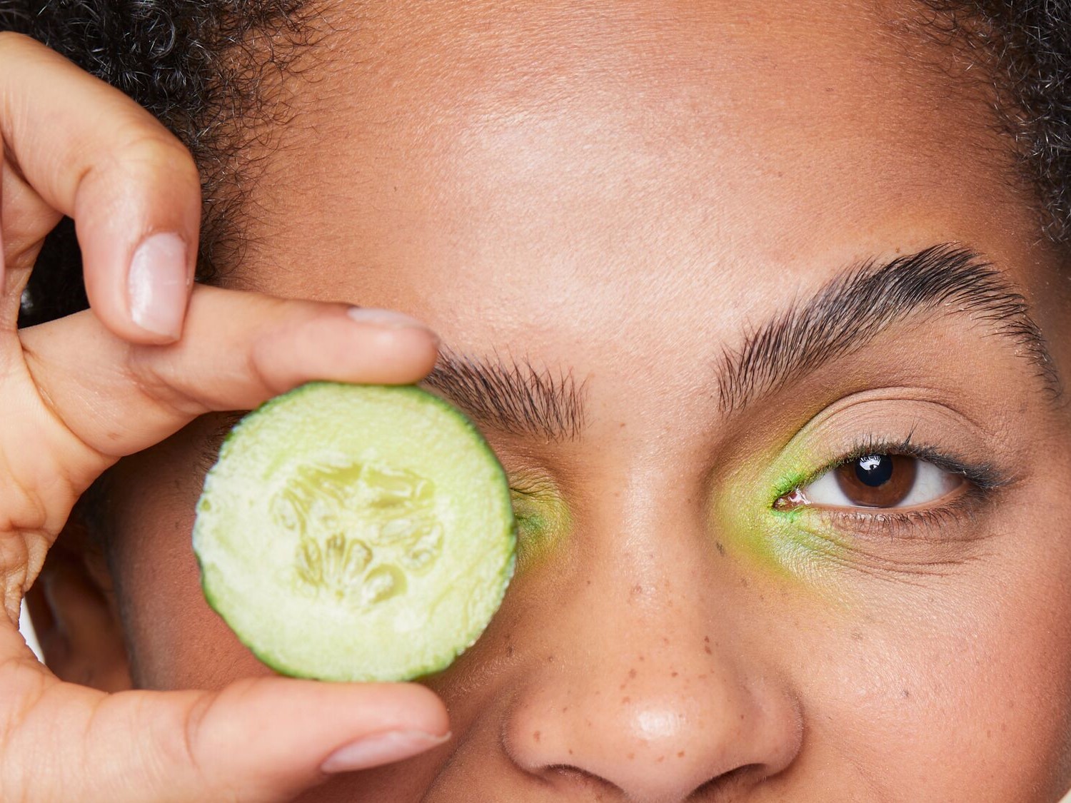 Nahaufnahme eines Gesichts, bei dem ein Auge mit einer Gurkenscheibe bedeckt ist, während das andere Auge mit grünem Lidschatten geschminkt ist.