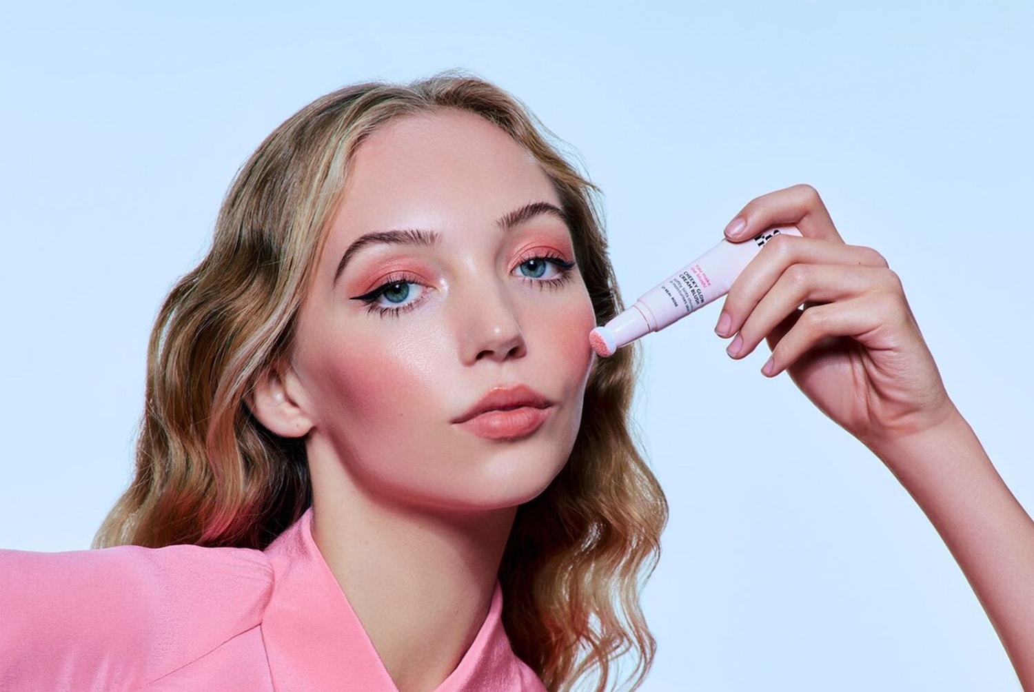 Een blonde vrouw met blauwe ogen brengt roze blush aan met een lichtroze tube Cheeky Glow Cream Blush, passend bij haar roze oogschaduw en blouse.