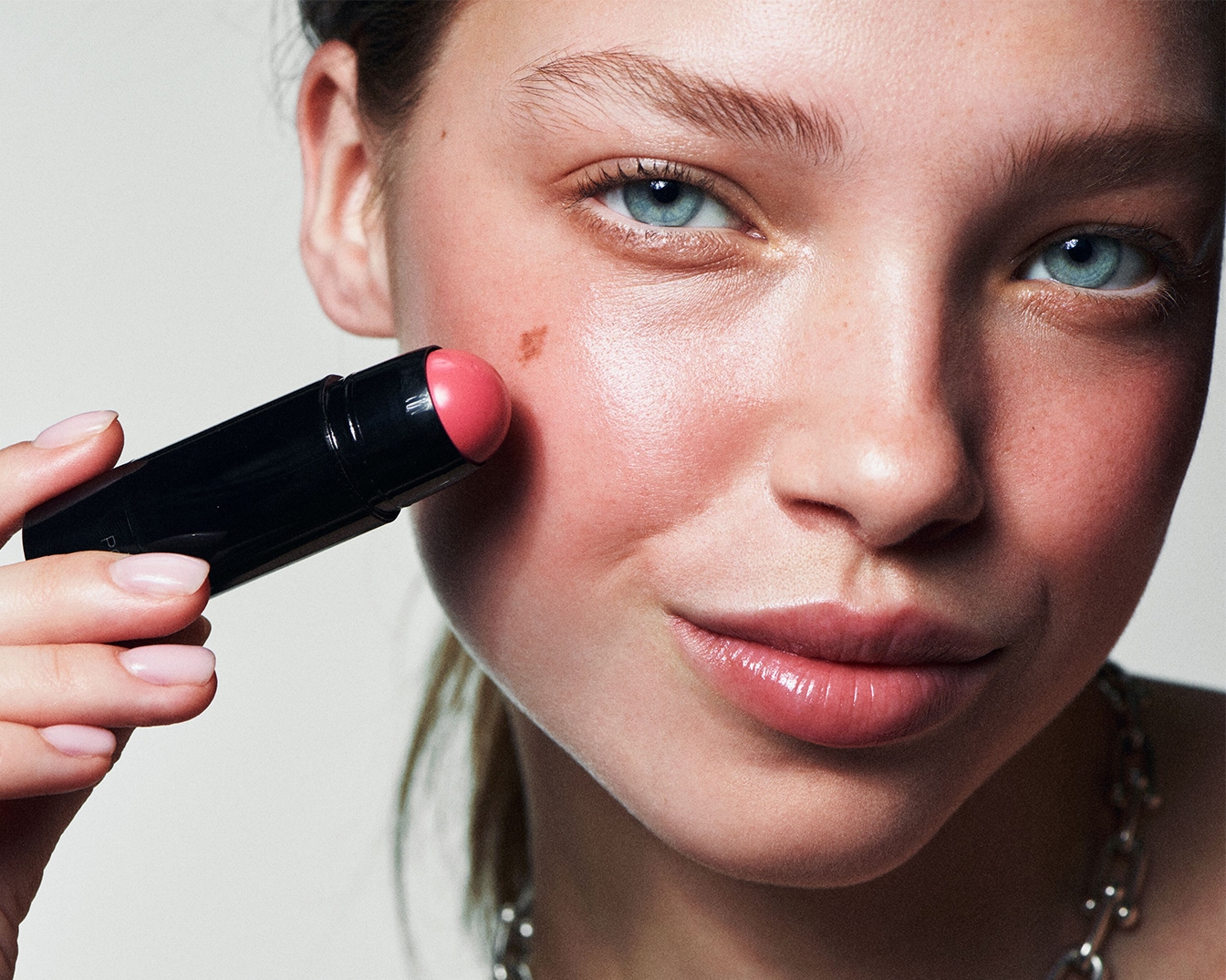 Une femme applique un blush rose crémeux sur sa joue avec un stick noir, pour un teint frais et lumineux.