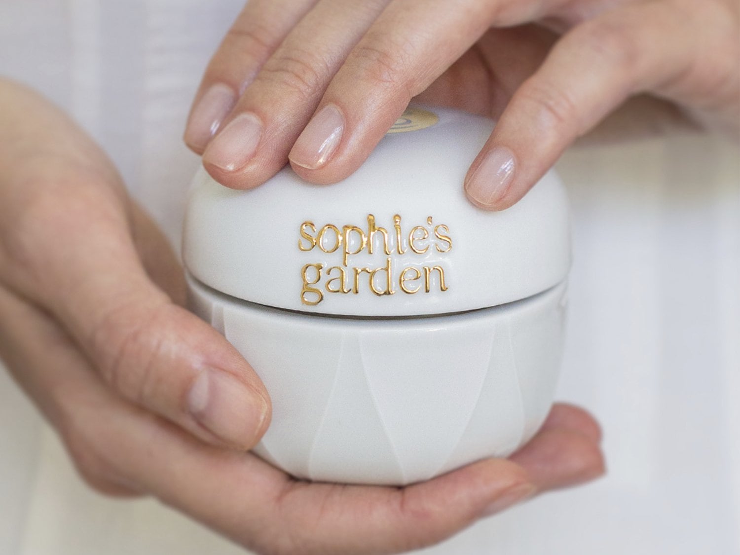 Primer plano de un tarro blanco de Sophie's Garden sostenido por dos manos, con el nombre de la marca en letras doradas.