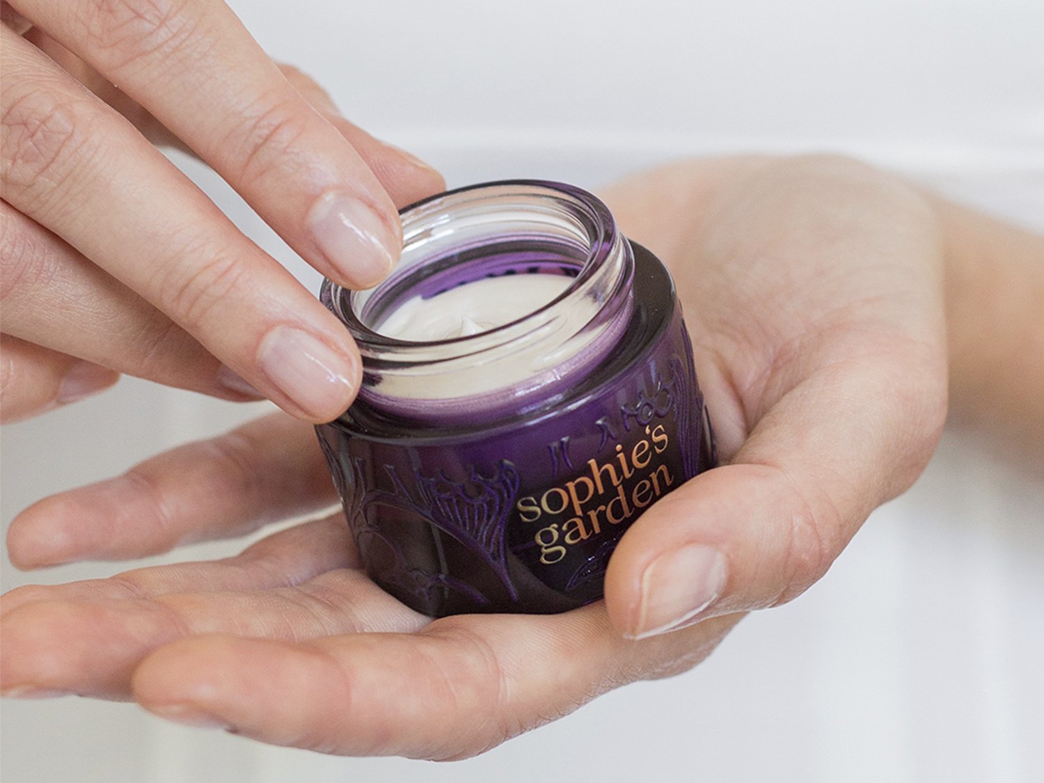 Primer plano de una mano sosteniendo un frasco morado de crema facial Sophie's Garden, con la tapa abierta, mostrando la crema blanca en su interior.