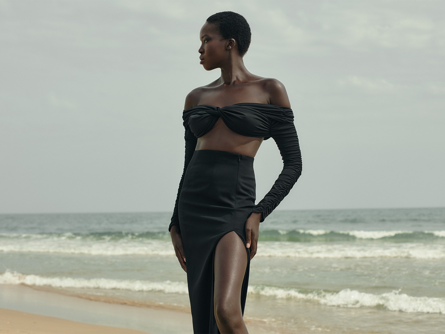Une femme noire élégante, vêtue d'un ensemble noir deux pièces avec un haut bandeau et une jupe fendue, pose sur une plage au bord de l'océan.