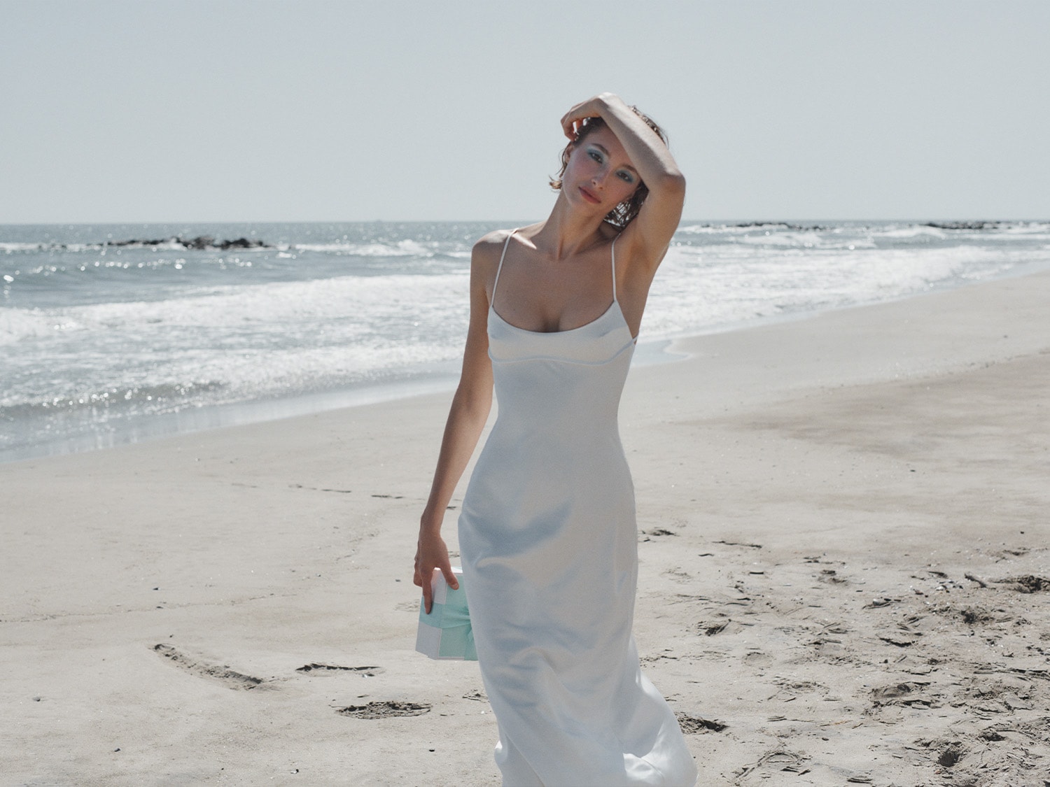 Une femme sur la plage, portant une robe blanche fluide et tenant une boîte turquoise, avec l'océan en arrière-plan.