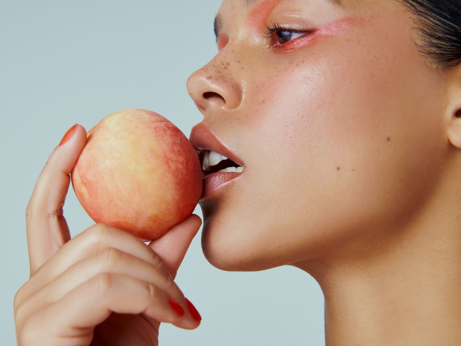 Nahaufnahme einer Frau mit orangefarbenem Lidschatten und roten Nägeln, die genüsslich in einen Pfirsich beißt.