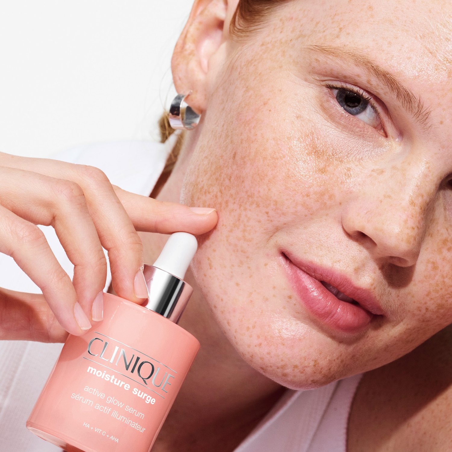 Nahaufnahme einer Frau mit Sommersprossen, die Clinique Moisture Surge Active Glow Serum in einer lachsfarbenen Flasche mit silbernem Verschluss und Pipette hält.