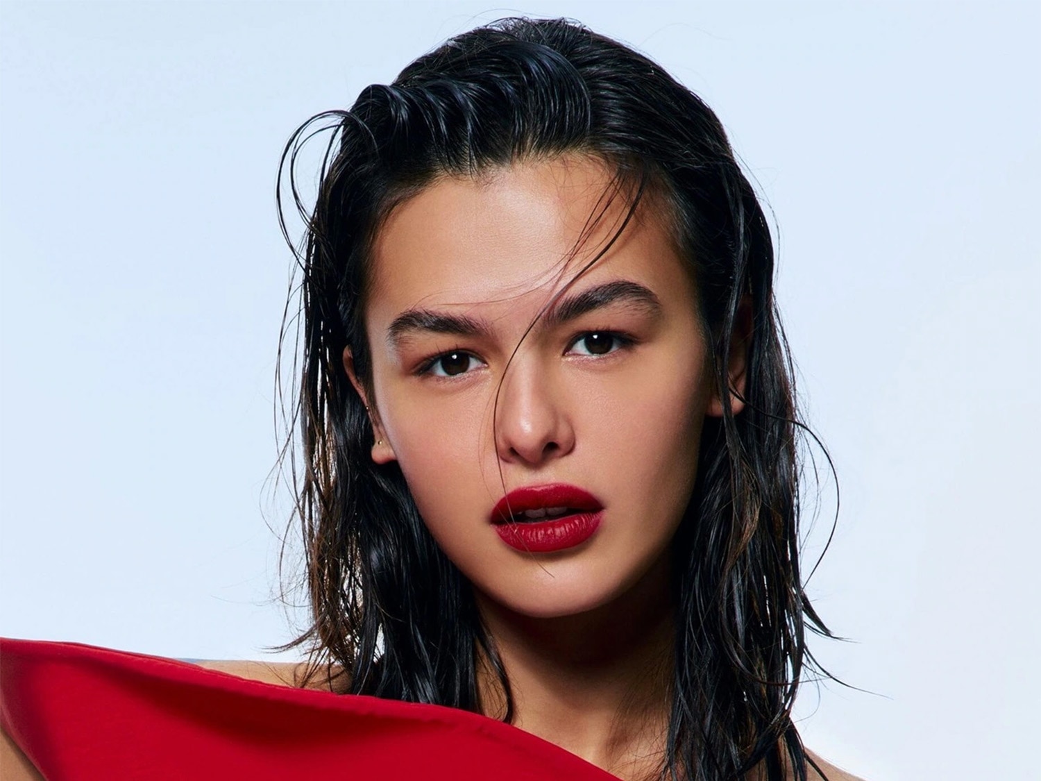 Portret van een vrouw met nat, donker haar en dieprode lippenstift, gekleed in een rode top tegen een heldere achtergrond.