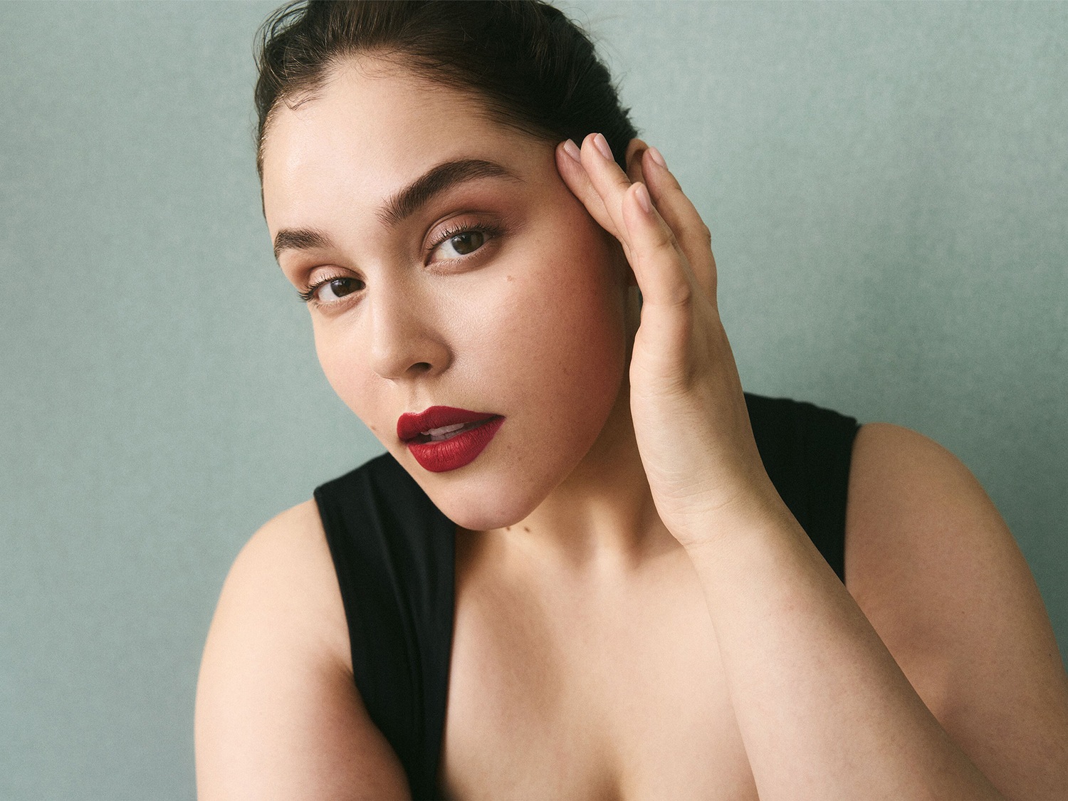 Een vrouw met een zwarte top en rode lippenstift, haar hand rustend op haar gezicht, tegen een zachte, effen achtergrond.