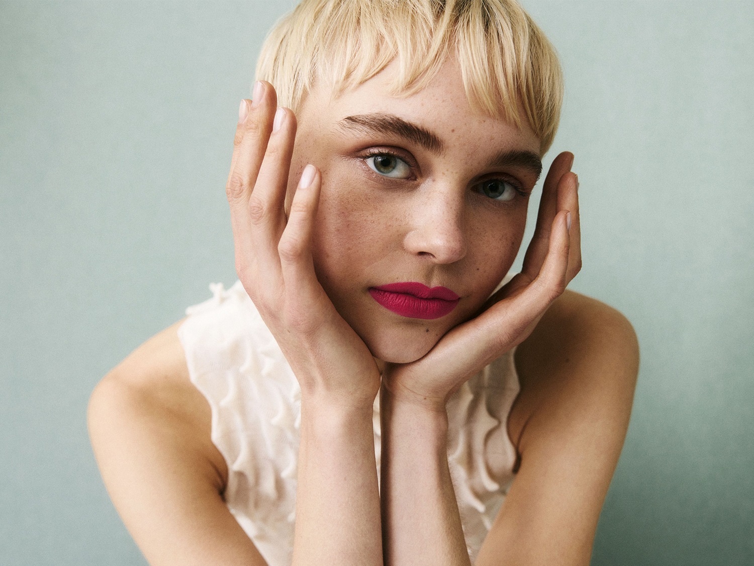 Portret van een jonge vrouw met blond haar en sproeten, die haar gezicht omlijst met haar handen en opvallende roze lippenstift draagt.