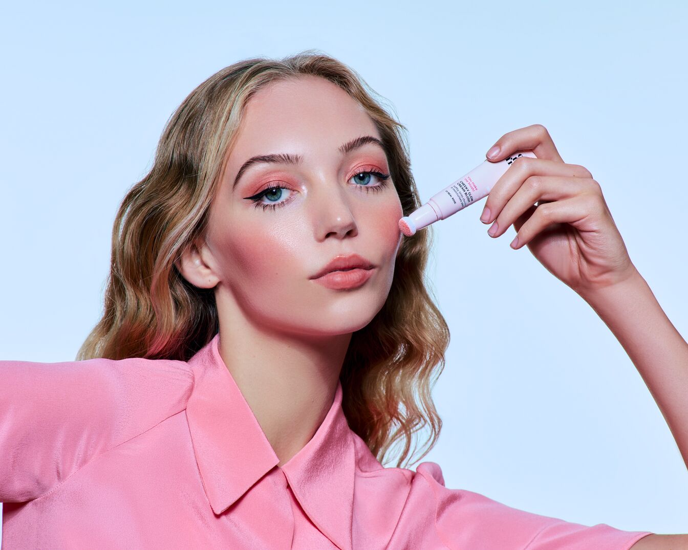 Eine junge Frau mit blonden Haaren und einem pinkfarbenen Hemd hält einen hellrosa Creme-Rouge-Stift an ihre Wange, der einen rosigen Schimmer auf ihrer Haut hinterlässt.