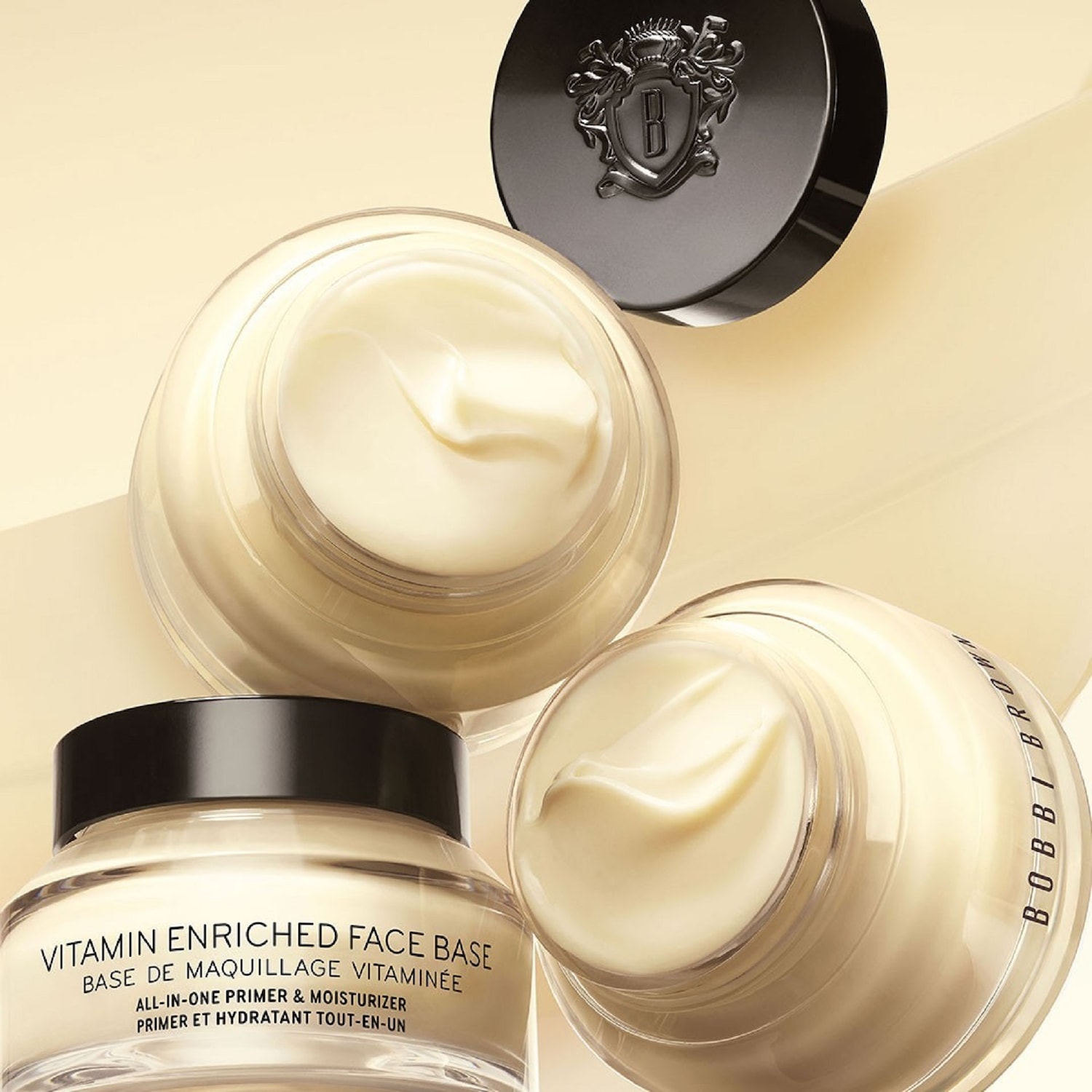 Kremowa baza pod makijaż Bobbi Brown Vitamin Enriched Face Base w przezroczystym słoiczku z czarną nakrętką, obok leży czarna nakrętka z logo marki.