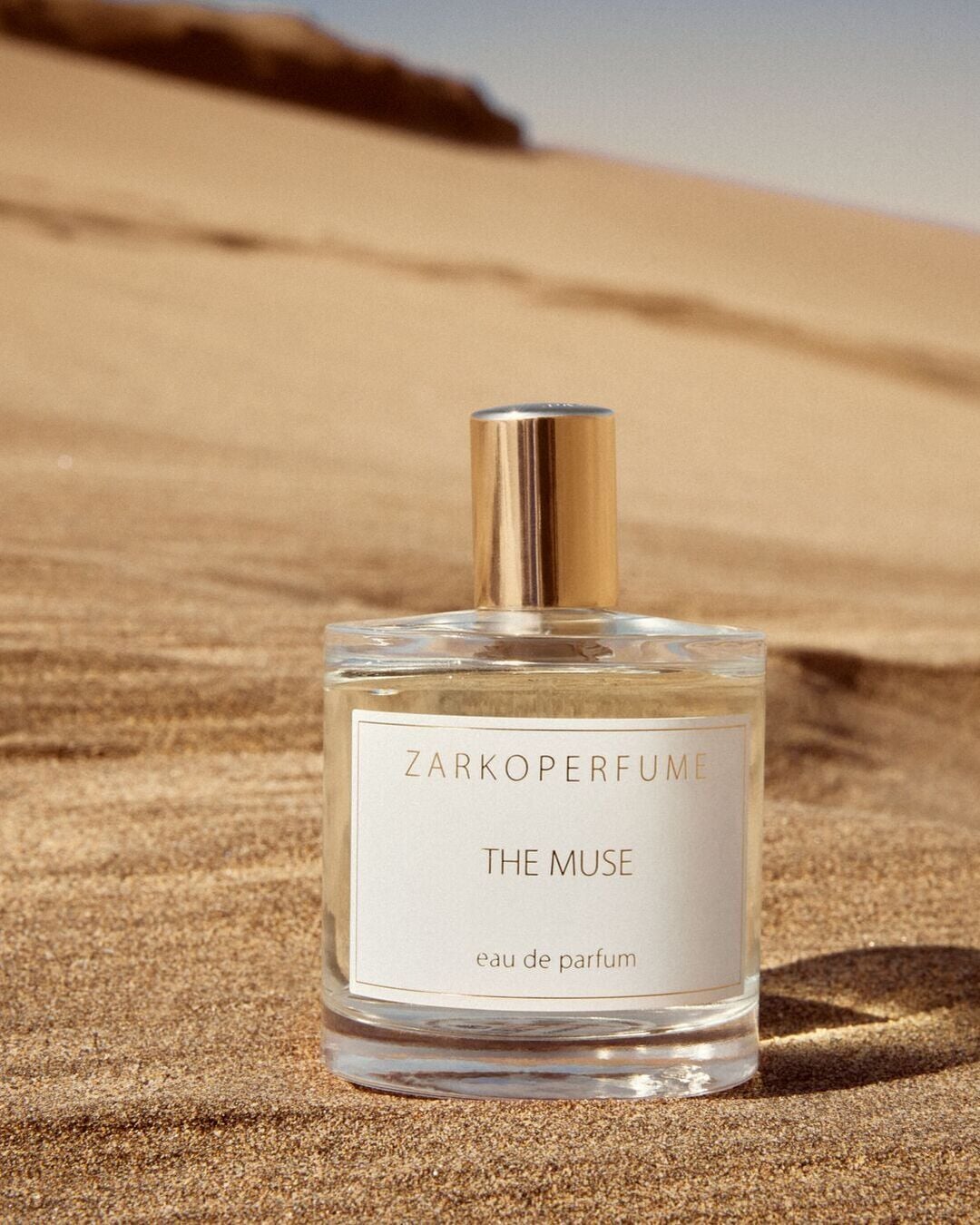 Smėlio fone pavaizduotas skaidraus stiklo „Zarkoperfume The Muse“ buteliukas su auksiniu dangteliu, suteikiantis elegancijos ir rafinuotumo įspūdį.
