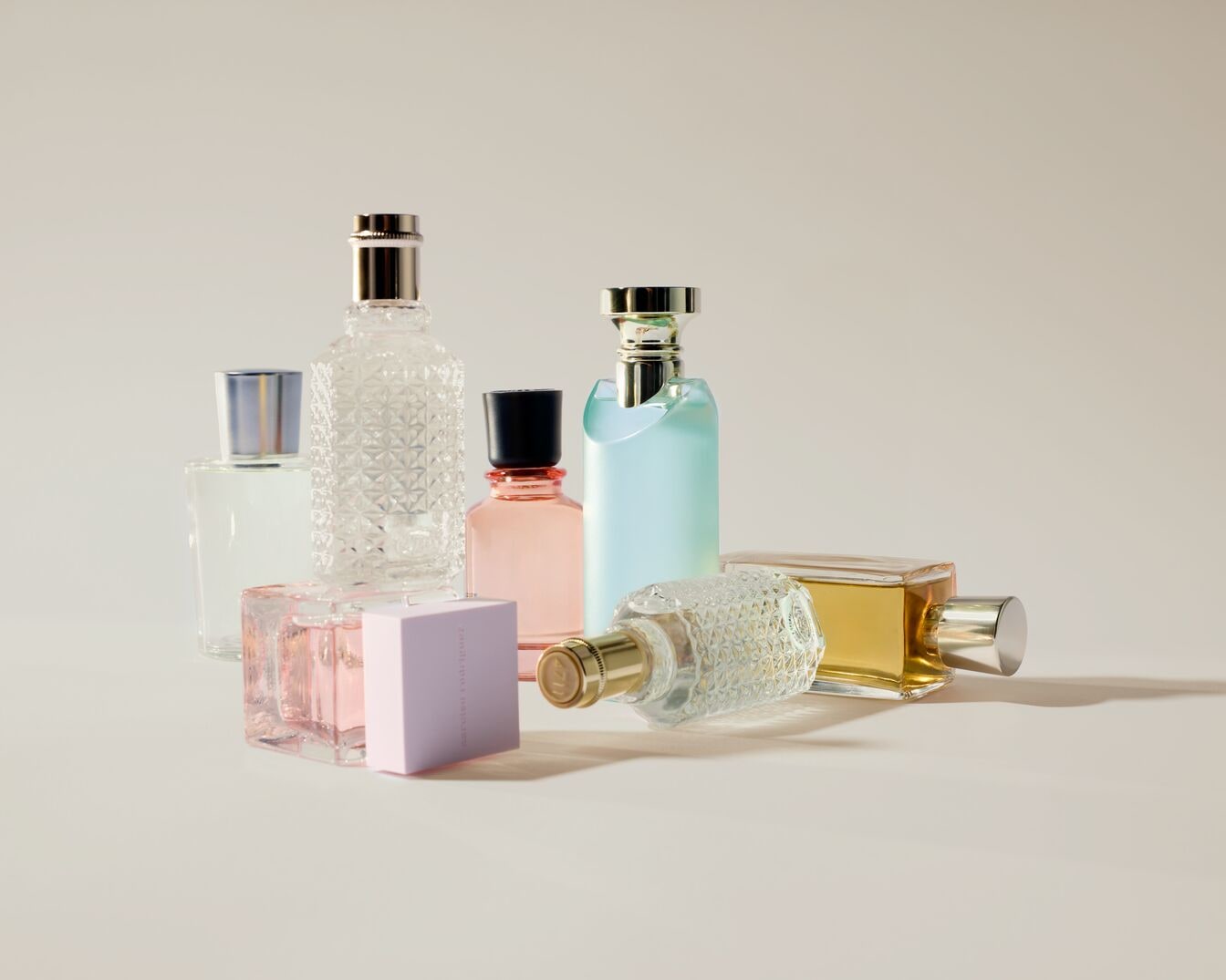 Įvairių formų ir spalvų kvepalų buteliukų kolekcija, įskaitant skaidrius, rožinius, žydrus ir auksinius atspalvius, išdėstytus ant švelnaus fono.