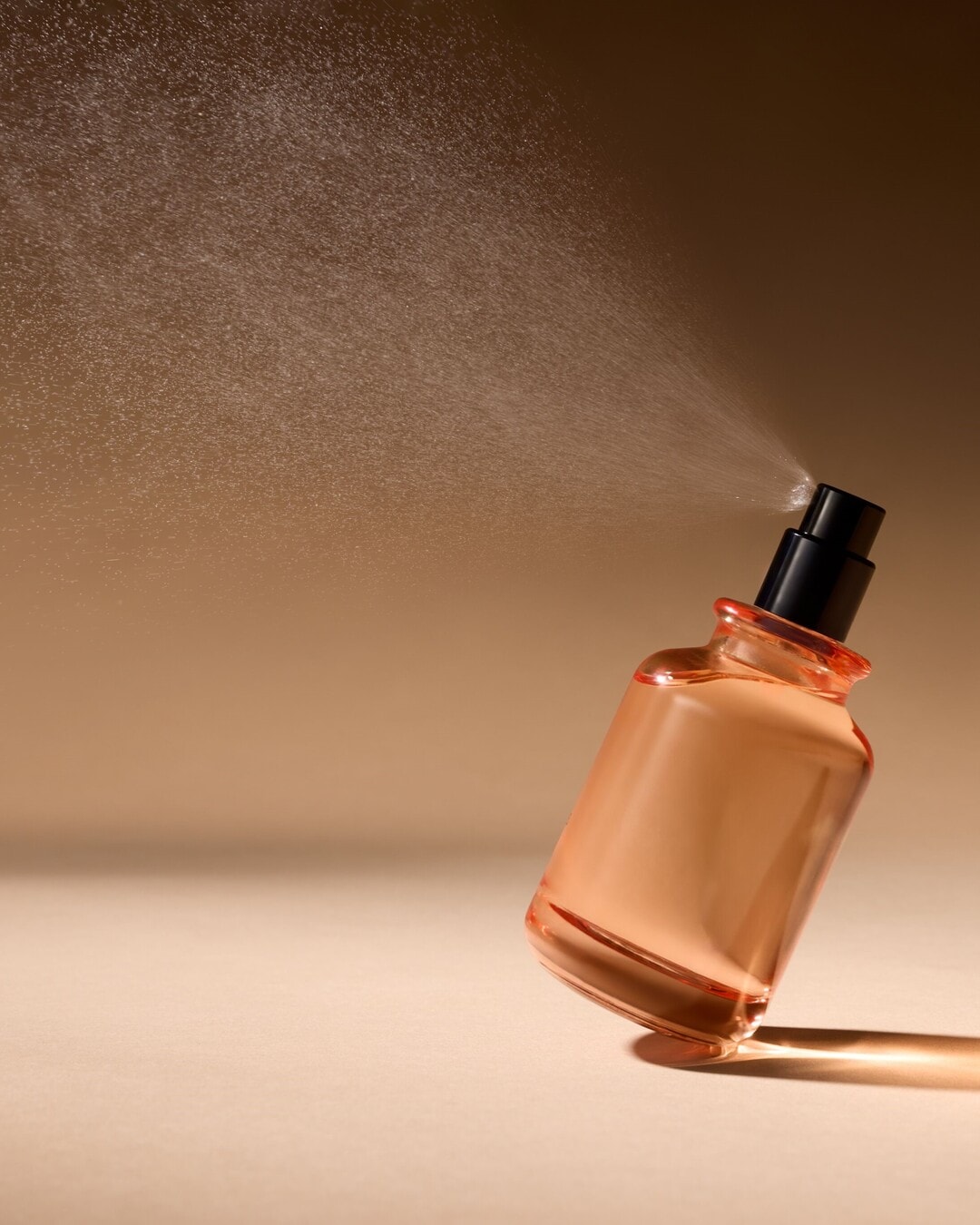 Persiko spalvos stiklinis kvepalų buteliukas su juodu purkštuvu purškia smulkią dulksną priešais smėlio spalvos foną.