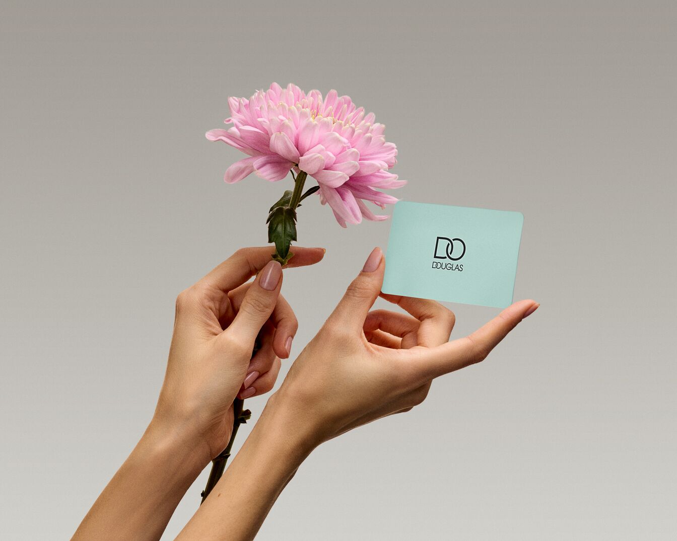 Delikatne dłonie trzymają różowy kwiat i kartę podarunkową Douglas na tle szarości, symbolizując piękno i możliwość obdarowania bliskich.