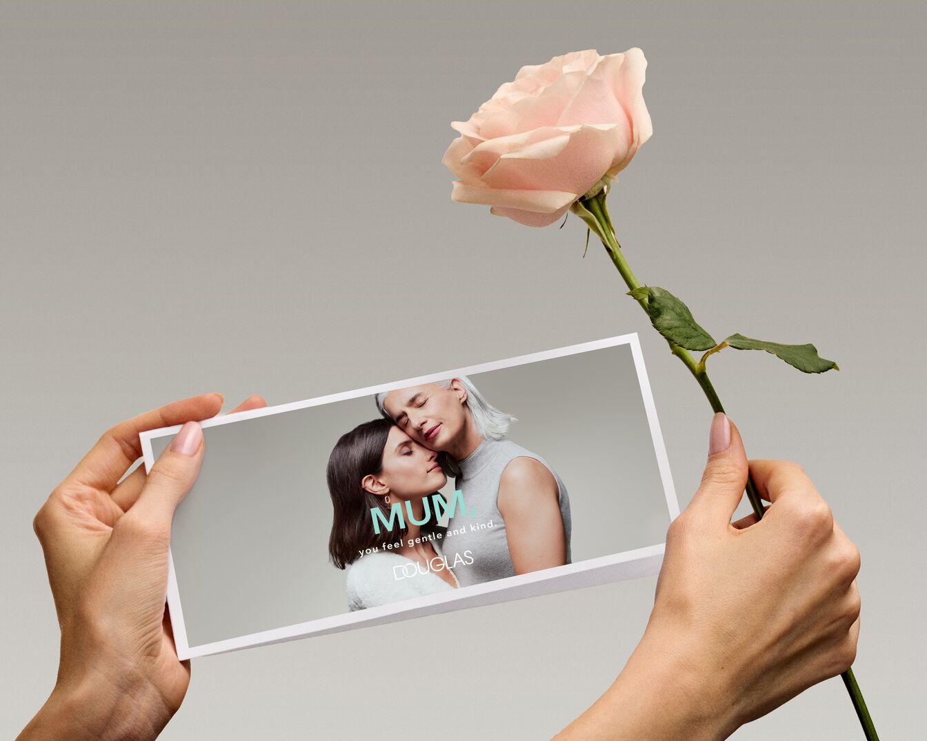 Na zdjęciu dłoń trzymająca kartkę z napisem „MUM you feel gentle and kind DOUGLAS” oraz wizerunkiem kobiety i starszej kobiety, obok znajduje się różowa róża.